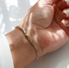 A delicate gold chain bracelet with a curb link design and a spring ring clasp, shown worn on a person's wrist. The hand has white manicured nails, and the background is a clean white surface with soft, natural shadows.