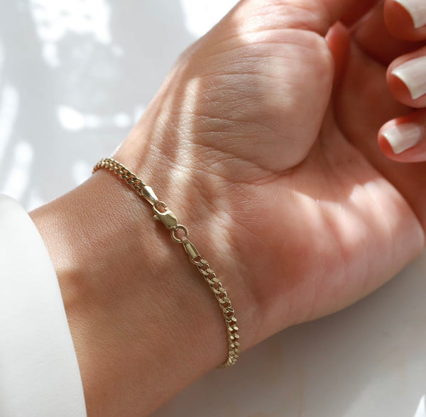 A delicate gold chain bracelet with a curb link design and a spring ring clasp, shown worn on a person's wrist. The hand has white manicured nails, and the background is a clean white surface with soft, natural shadows.