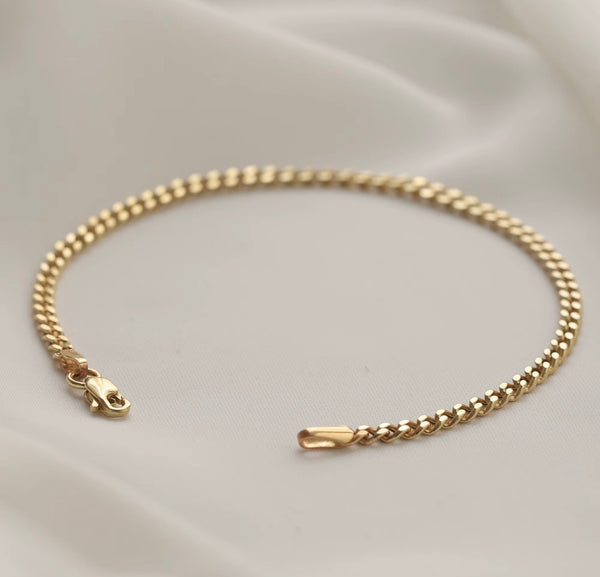 A gold-toned chain bracelet with a lobster clasp and a polished end tab, displayed on a soft white satin fabric background.